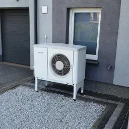 Biała jednostka zewnętrzna urządzenia grzewczego marki Stiebel Eltron na tle elewacji domu z oknem, umieszczona na żwirowym podłożu.