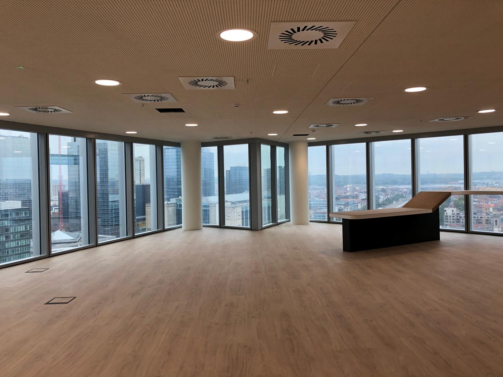 Wnętrze nowoczesnego biura z panoramicznymi oknami ukazującymi widok na miasto, podłoga wyłożona panelami drewnopodobnymi, sufit z perforowanej płyty z wbudowanymi okrągłymi lampami i kratkami...