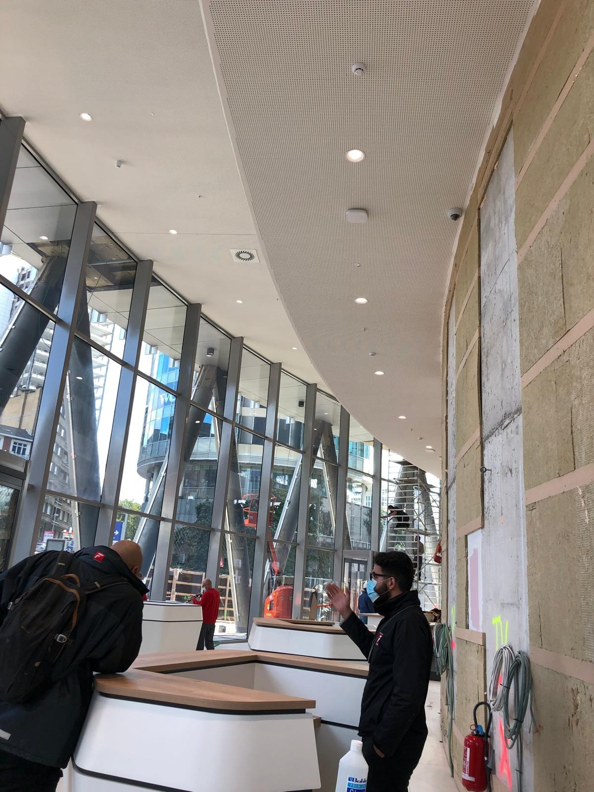 Wnętrze budynku w trakcie wykańczania, widoczne ocieplone ściany z wełny mineralnej, elementy konstrukcji stalowej i szklana fasada, pracownicy w tle.