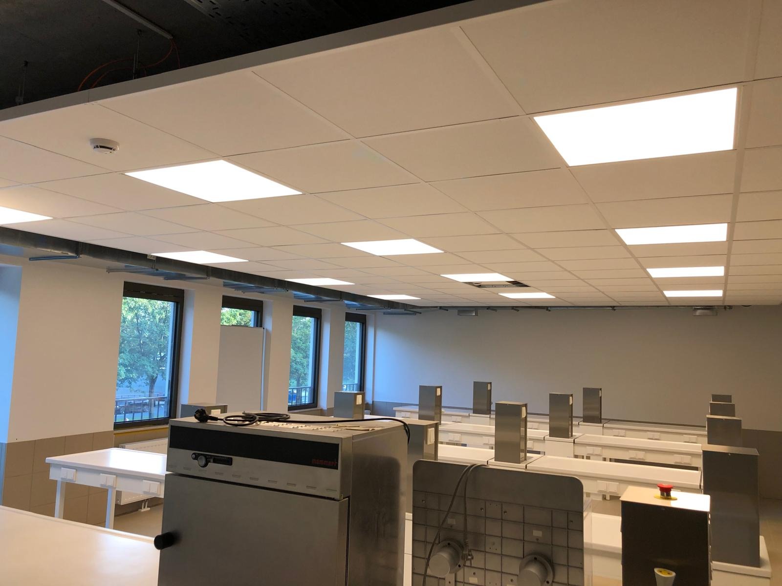 Nowoczesne oświetlenie sufitowe w przestronnym, jasnym pomieszczeniu z białymi ścianami i rzędami metalowych stanowisk pracy, widoczne okna z zielenią na zewnątrz.