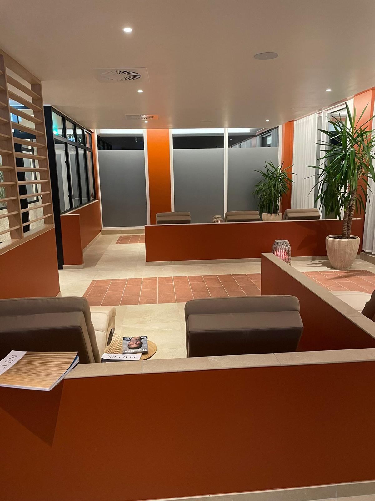 Nowoczesne wnętrze poczekalni z pomarańczowymi ścianami, szarymi szybami, beżową podłogą z pomarańczowymi akcentami, roślinami doniczkowymi i brązowymi sofami. Na stoliku leżą czasopisma.