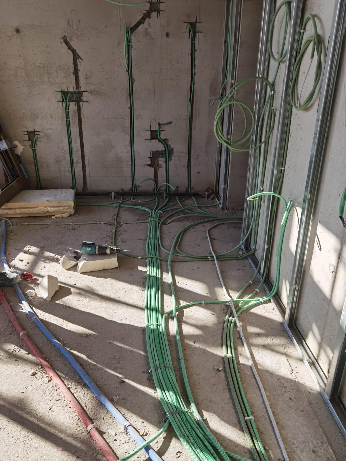 Wnętrze w trakcie instalacji elektrycznej: zielone rury karbowane prowadzone po surowych ścianach i podłodze, widoczne metalowe profile ścian działowych, w tle narzędzie - wiertarka akumulatorowa.