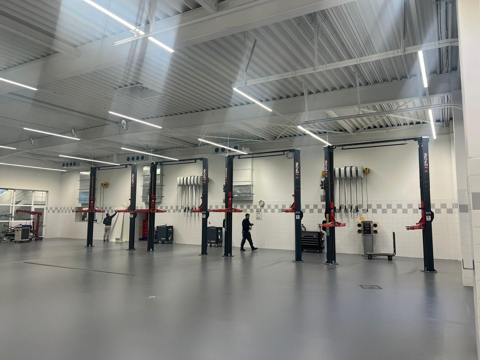 Wnętrze nowo wybudowanego warsztatu samochodowego z kilkoma podnośnikami dwukolumnowymi, białymi ścianami wyłożonymi częściowo kafelkami i szarą posadzką epoksydową.