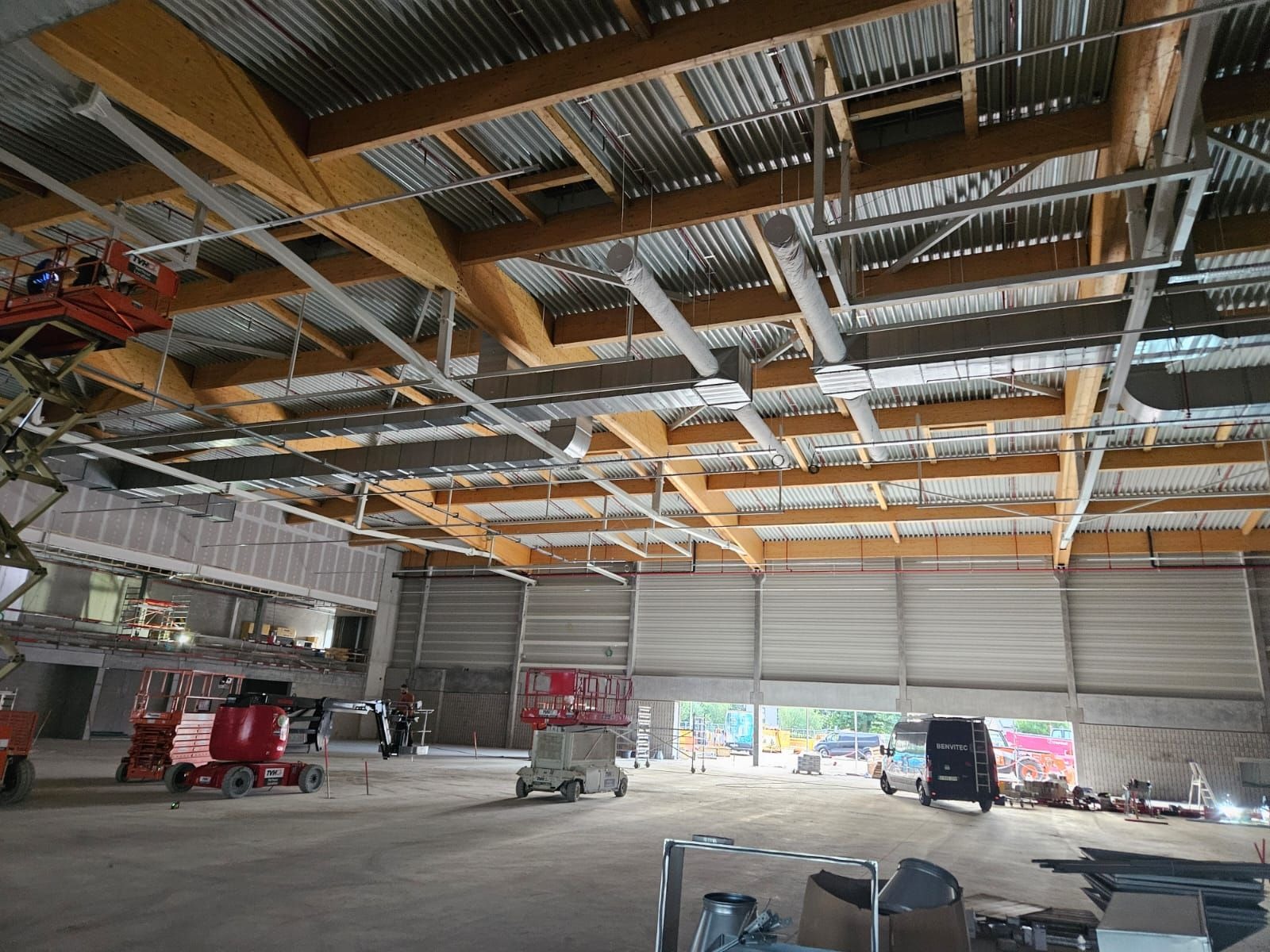 Wnętrze hali przemysłowej w trakcie budowy z widoczną drewnianą konstrukcją dachu, metalową blachą falistą, instalacją wentylacyjną i podnośnikami koszowymi.