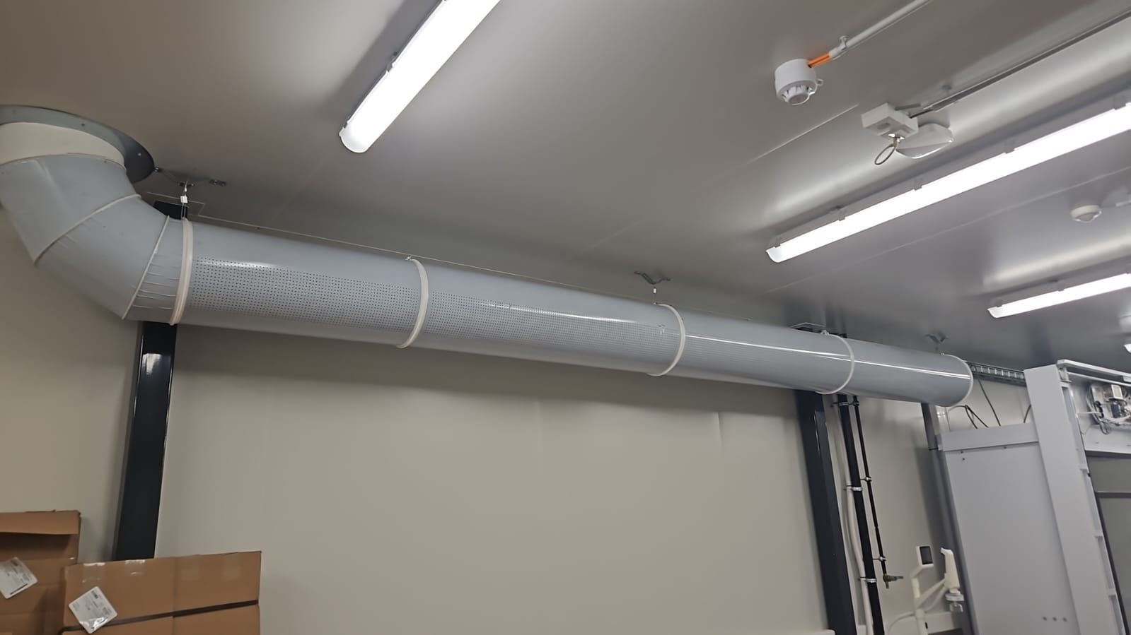 Szary, perforowany kanał wentylacyjny zamontowany pod sufitem pomieszczenia przemysłowego, widoczne elementy mocujące i oświetlenie LED.