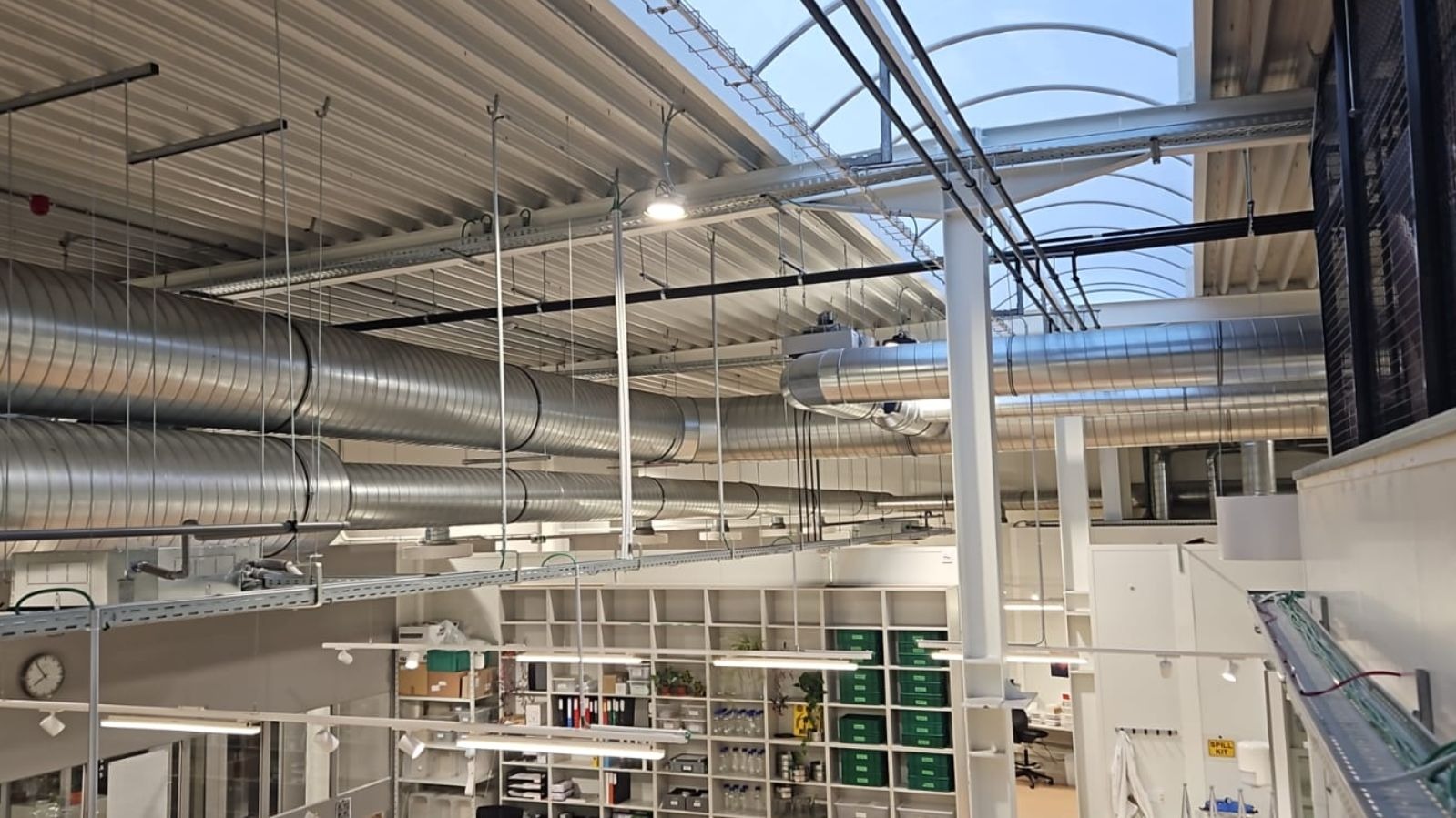 Wnętrze hali z widocznymi, spiralnymi kanałami wentylacyjnymi pod sufitem, oświetleniem LED i regałami z wyposażeniem laboratoryjnym w tle.