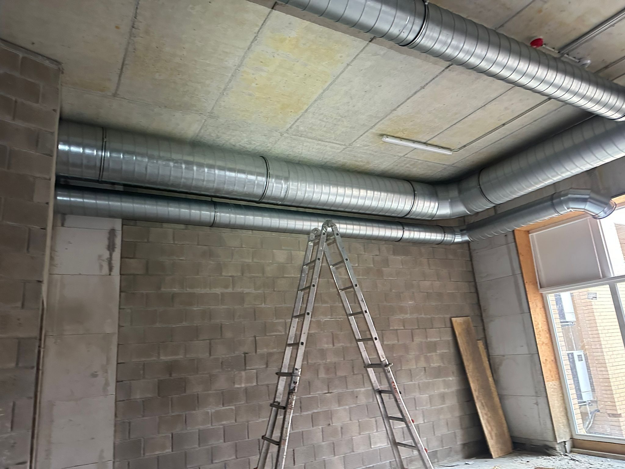 Montaż spiralnych kanałów wentylacyjnych z blachy ocynkowanej pod betonowym sufitem, widoczna drabina aluminiowa i surowe ściany z bloczków betonowych w trakcie budowy.
