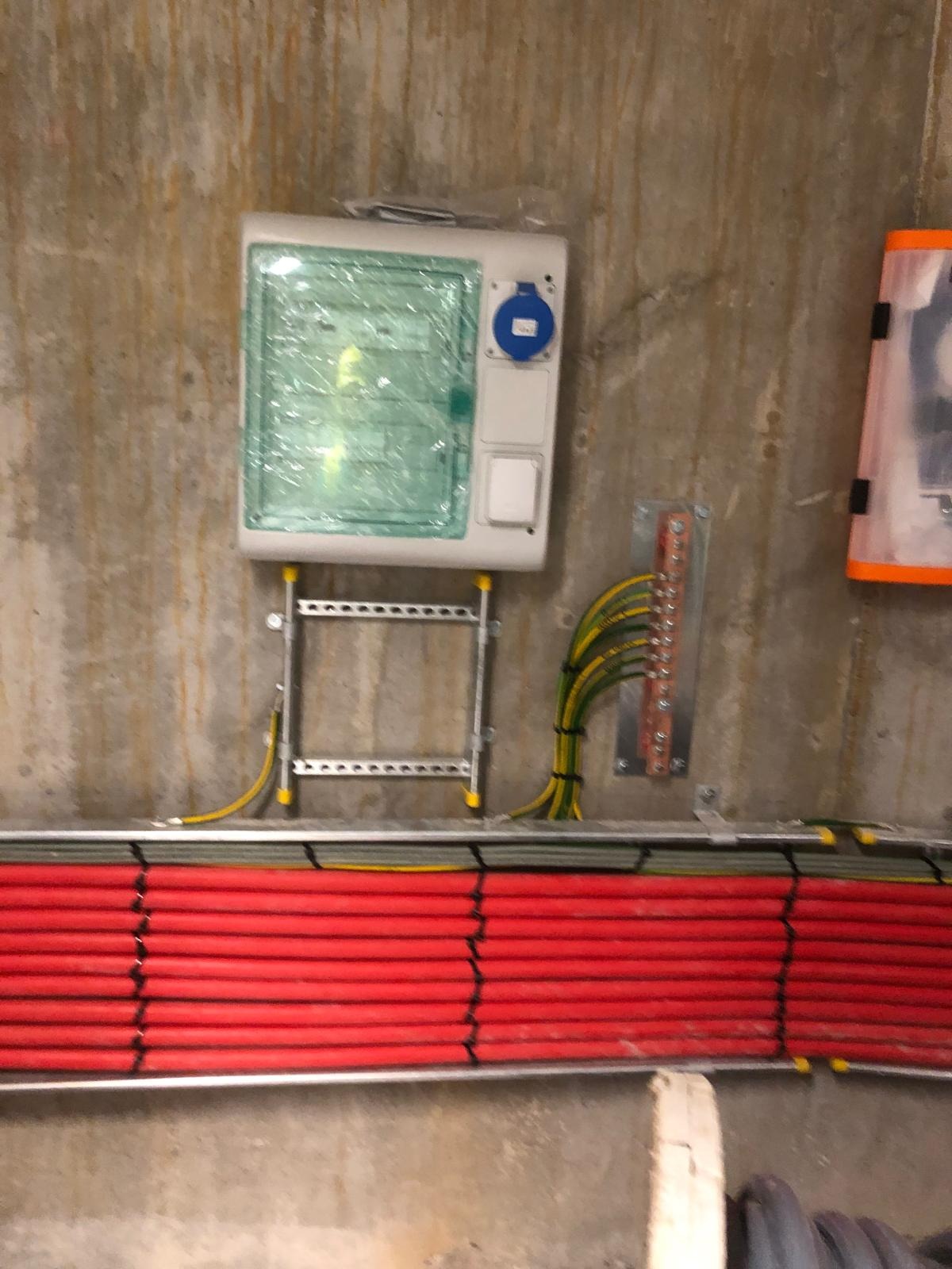 Nowa instalacja elektryczna w Korzennej: rozdzielnia, kable w czerwonych peszlach i listwa uziemiająca, montowane na surowej ścianie betonowej.