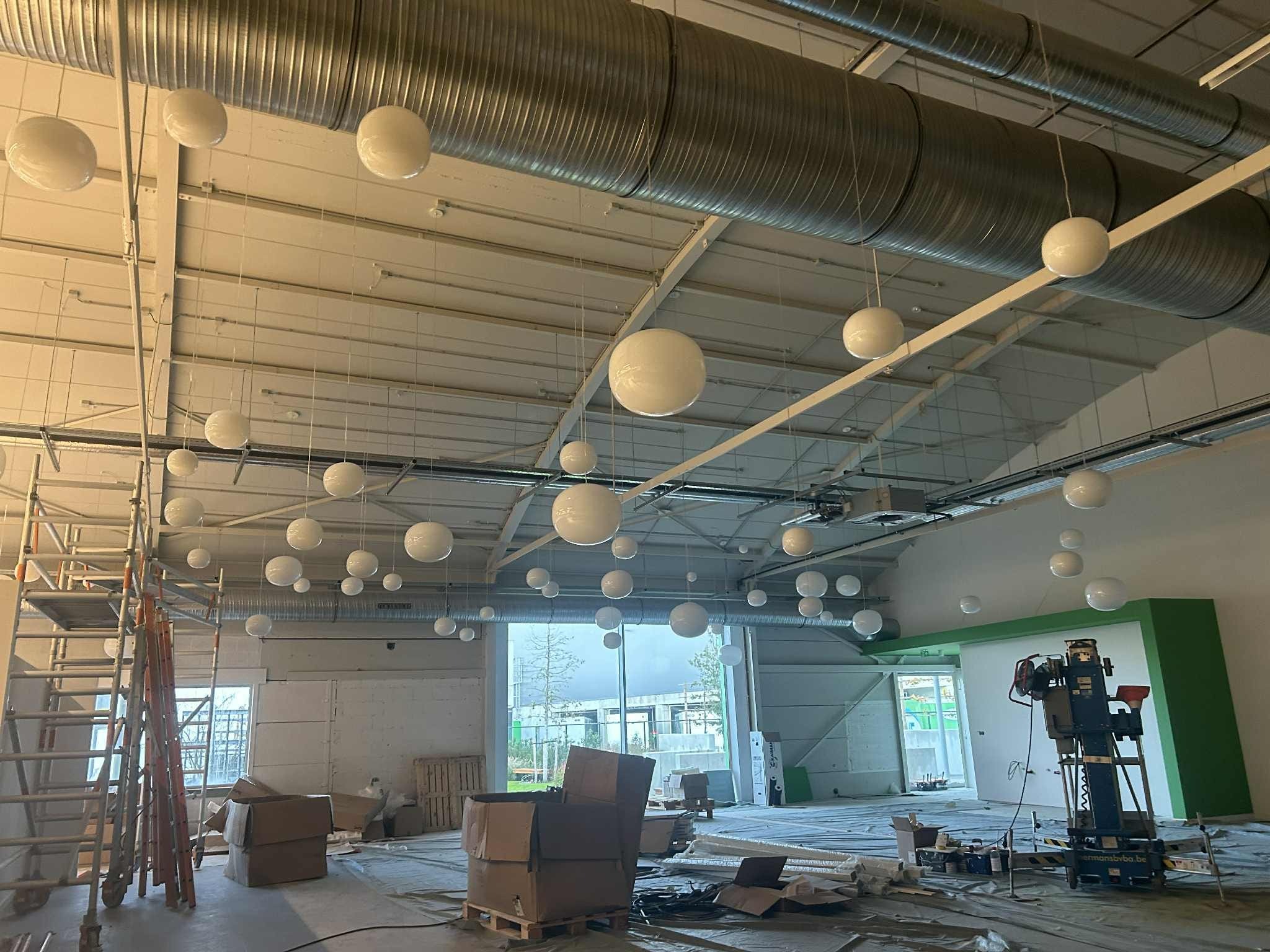 Instalacja oświetlenia w dużej hali: białe, kuliste lampy wiszą pod sufitem z widocznymi rurami wentylacyjnymi. Scaffolding i maszyna budowlana w tle.