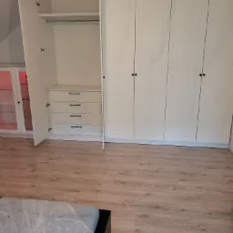 Biała zabudowa meblowa z szafami i szufladami, z otwartymi drzwiami do jednej z szaf, widoczny wieszak i półka, oświetlenie sufitowe w stylu industrialnym.