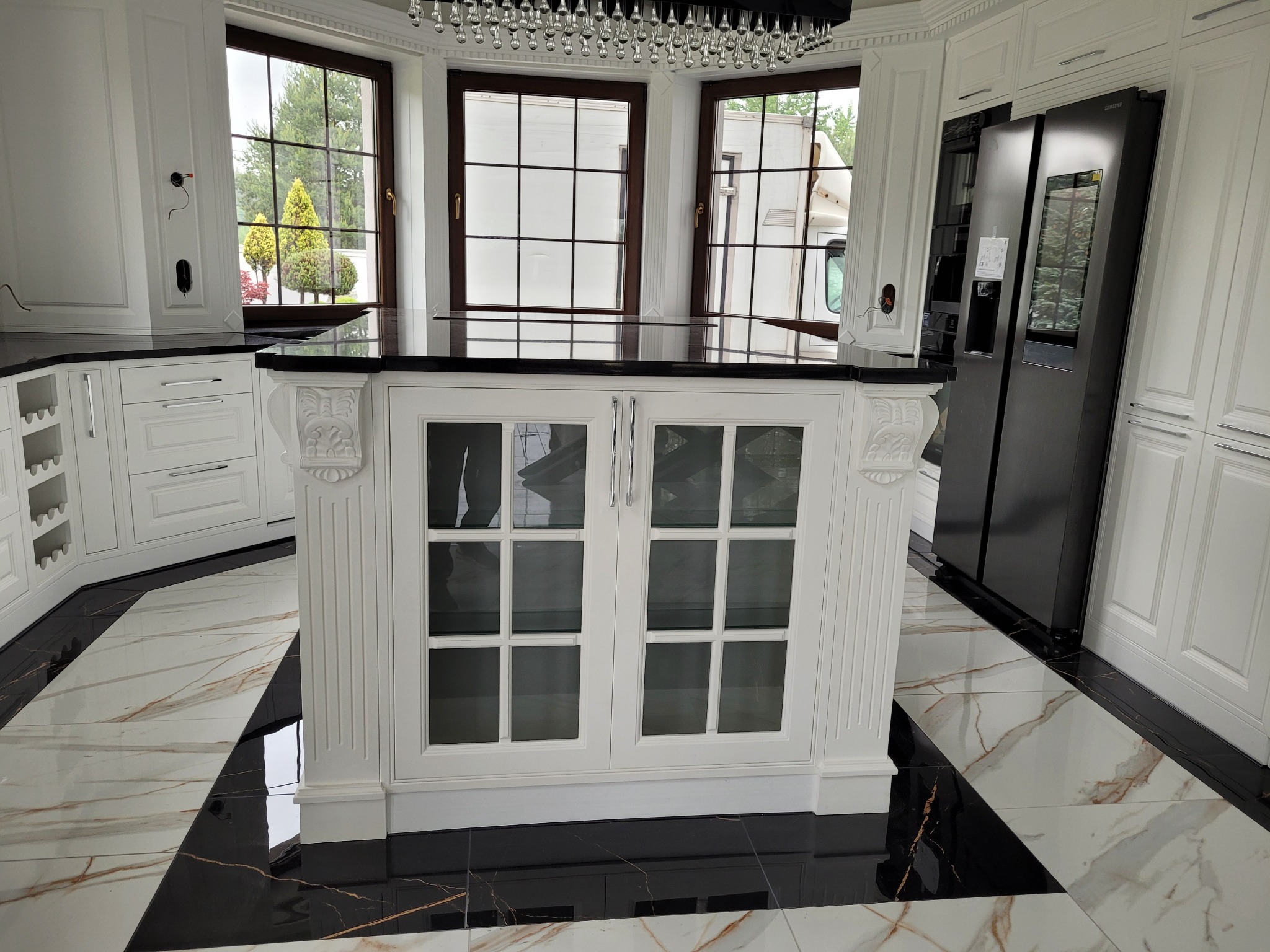 Nowoczesna kuchnia z wyspą, czarnym blatem i białymi, frezowanymi frontami szafek; podłoga w biało-czarne pasy, okna z widokiem na zieleń.