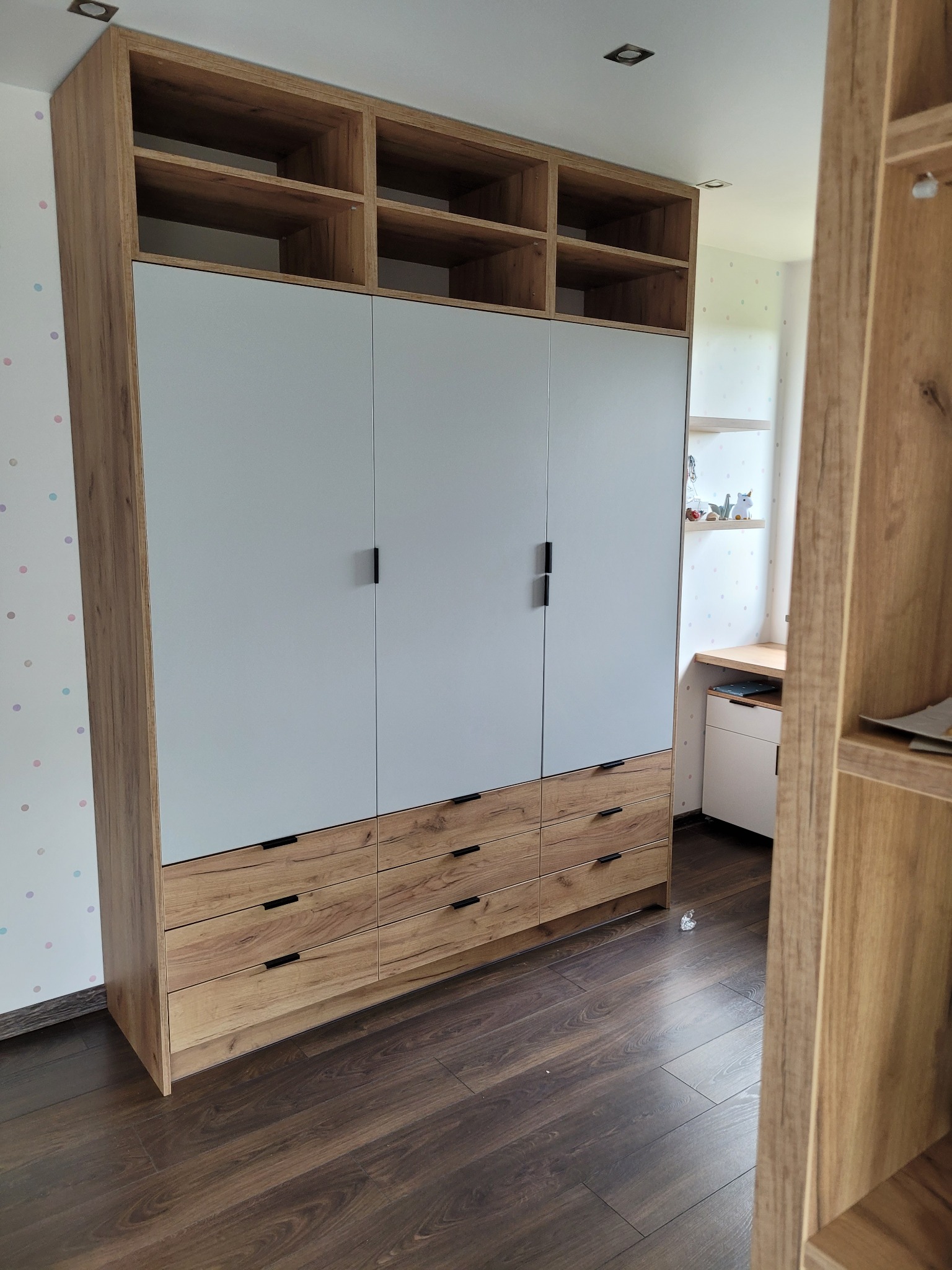 Wysoka szafa z szufladami i otwartymi półkami w górnej części, wykonana na zamówienie w Wadowicach, z białymi frontami i drewnopodobnymi elementami, widoczna na tle ściany w kropki.