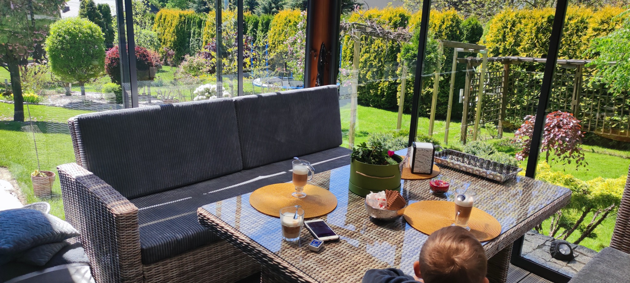 Wygodna sofa z szarej tkaniny w oranżerii, z widokiem na zadbany ogród, na stole kawa latte, ciasteczka i telefon.