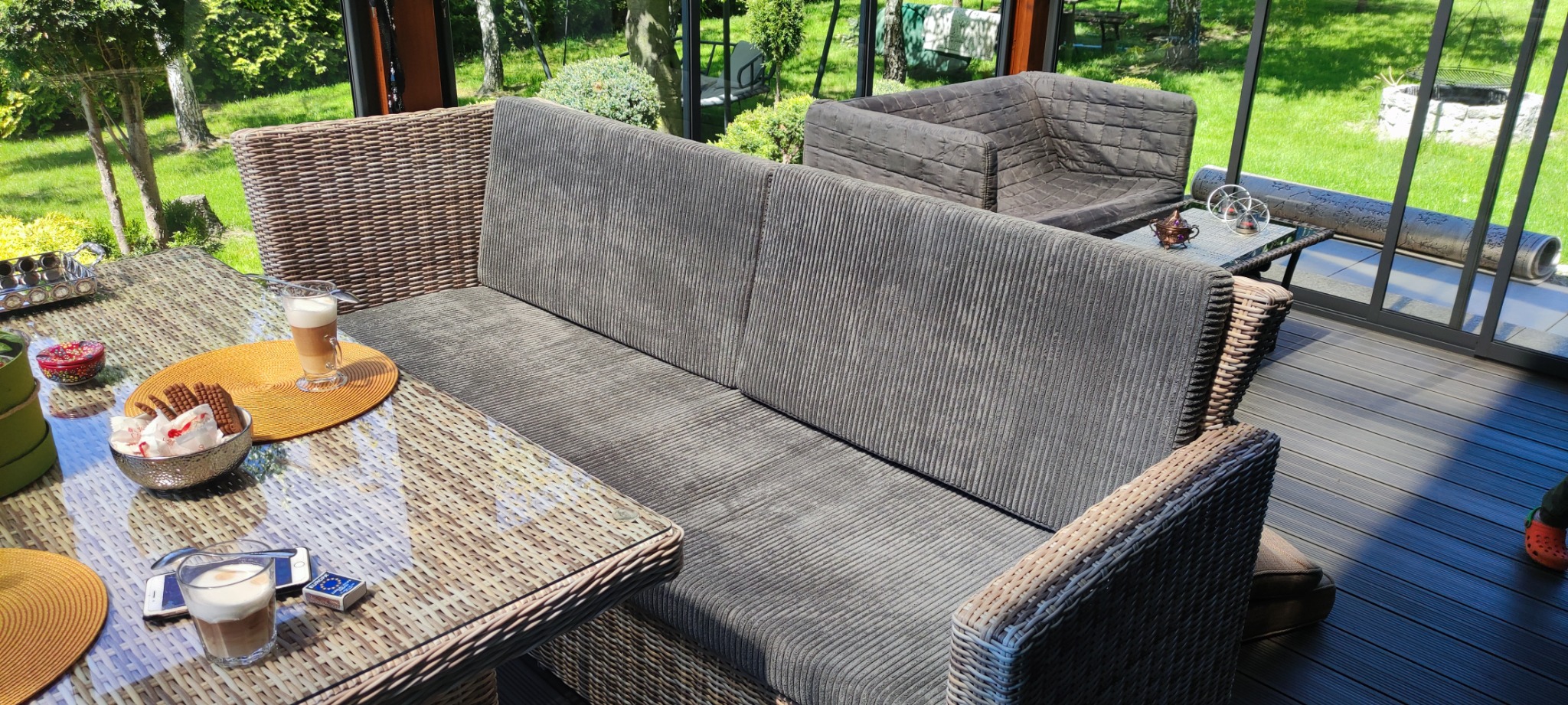 Zadaszony taras z rattanowymi meblami wypoczynkowymi: sofa z szarym, prążkowanym obiciem i stolik z napojami i deserami w słoneczny dzień.