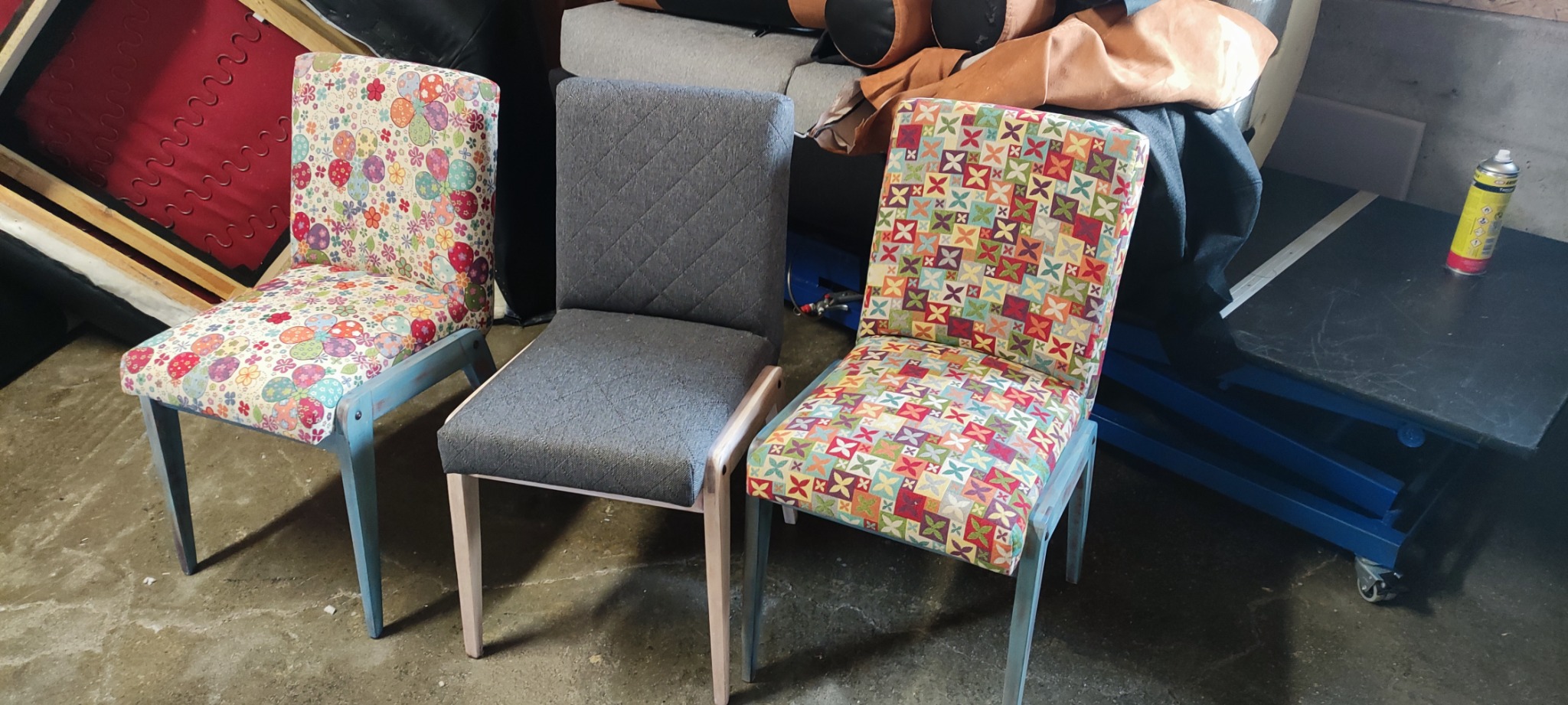 Trzy krzesła po renowacji tapicerskiej, każde z innym wzorem: kwiaty, geometryczny wzór w romby i patchworkowy wzór z kwadratów. Krzesła stoją w warsztacie, widoczne narzędzia i materiały w tle.