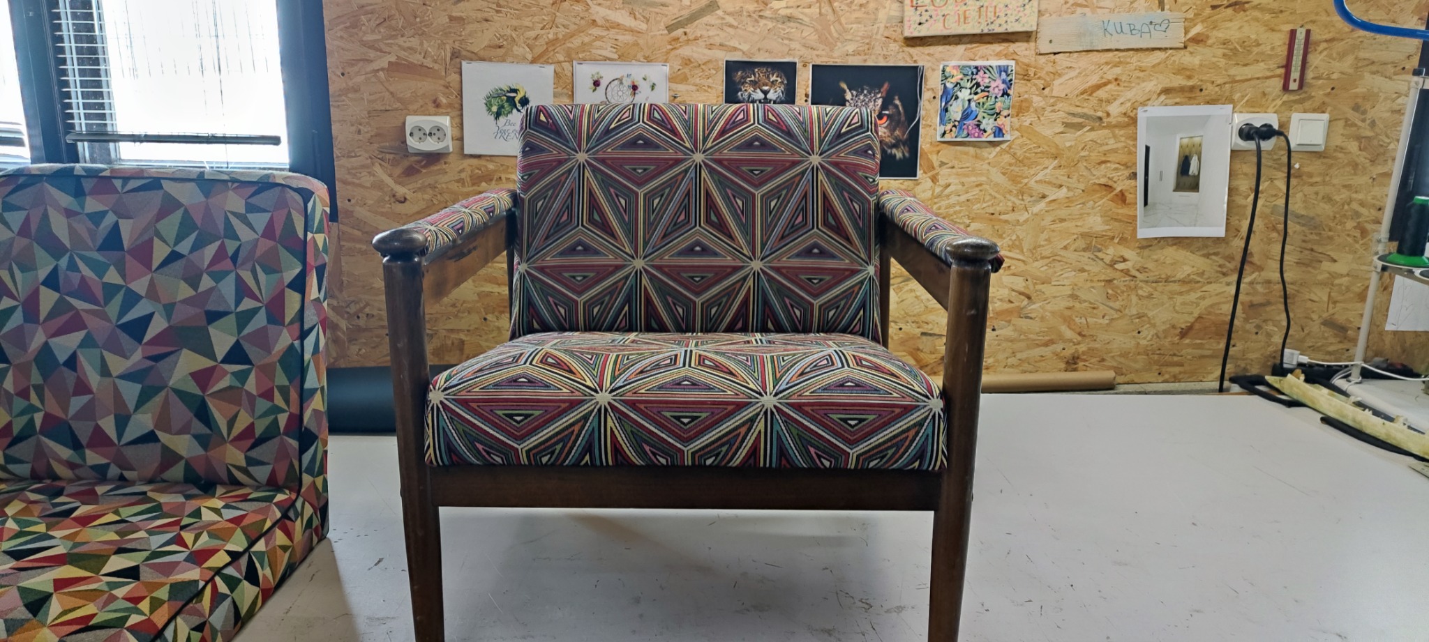 Odrestaurowany fotel z drewnianą ramą, obity tkaniną w geometryczne wzory w odcieniach czerwieni, zieleni i żółci, stojący w warsztacie z płyt OSB, na których wiszą obrazy i instalacje elektryczne.