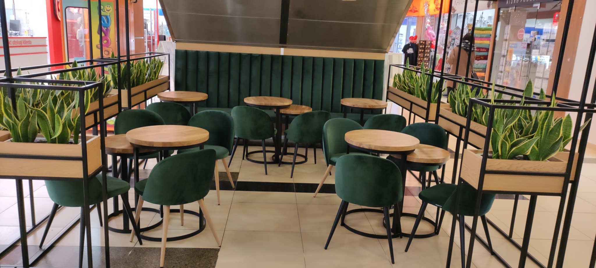 Zielone, tapicerowane krzesła z drewnianymi okrągłymi stolikami w przestrzeni handlowej, z roślinami w ozdobnych donicach
