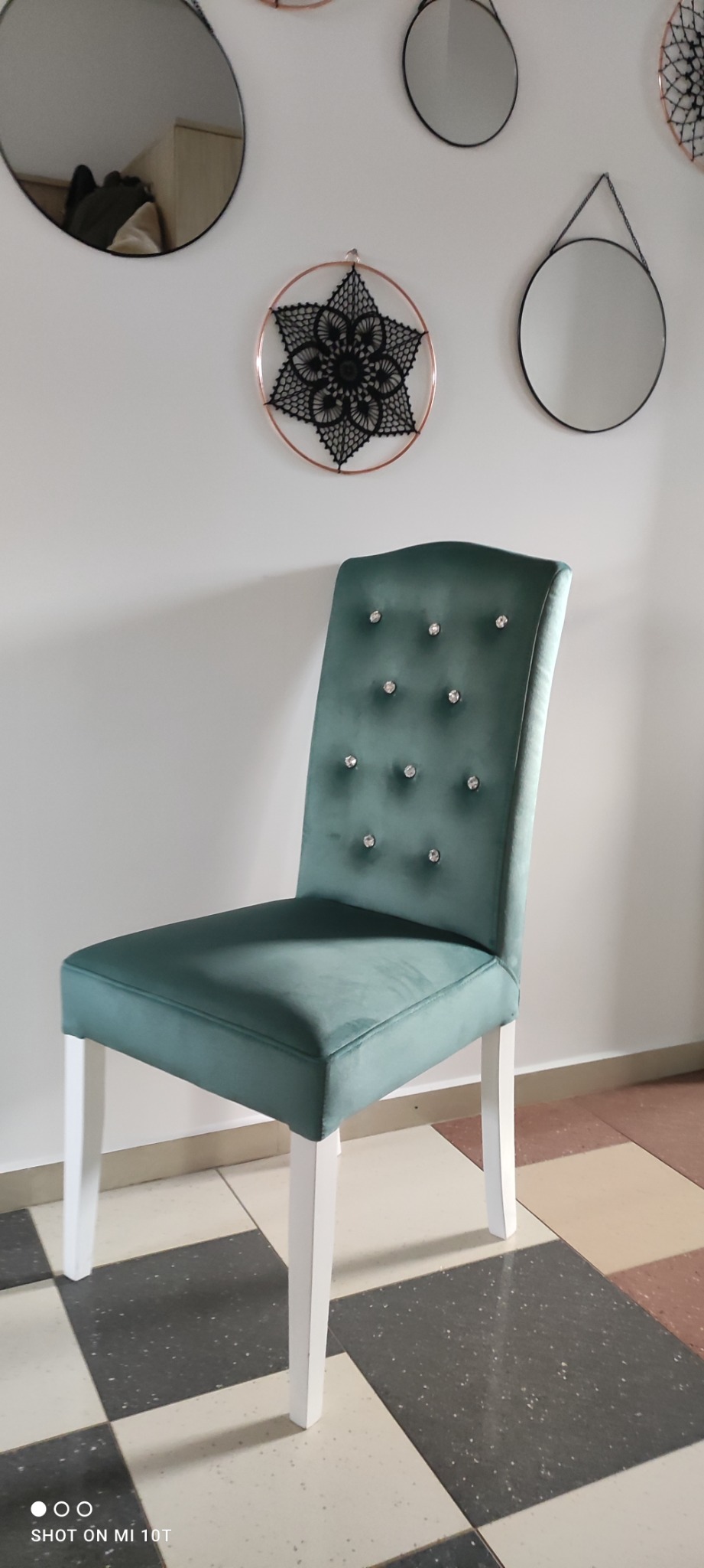 Krzesło tapicerowane zielonym welurem, z białymi nogami i dekoracyjnymi kryształkami na oparciu, na tle białej ściany z okrągłymi lustrami i ozdobą w stylu makramy.
