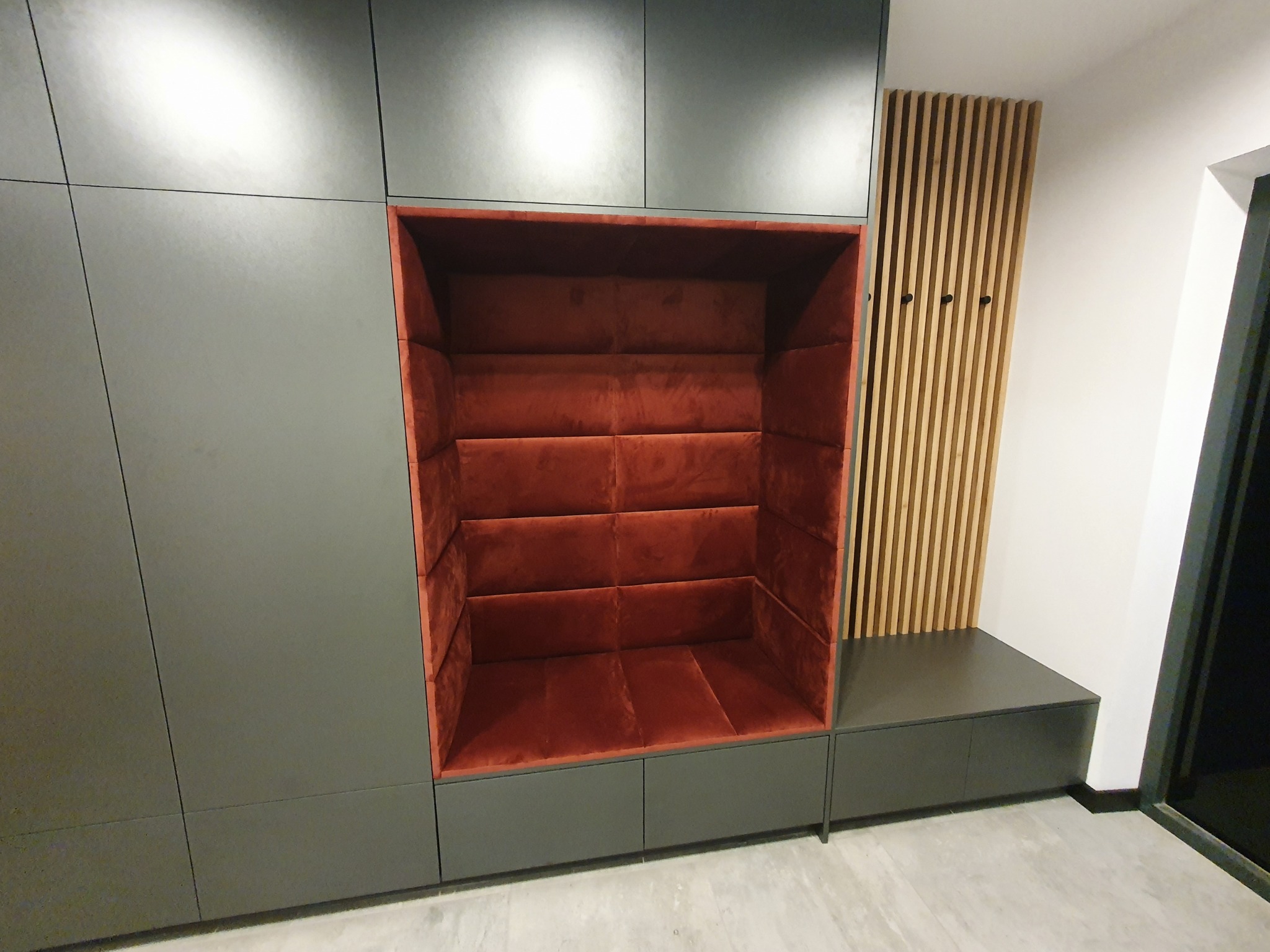 Wnęka z tapicerowanym, bordowym wnętrzem wbudowana w szarą zabudowę meblową, obok drewniany panel dekoracyjny z wieszakami.