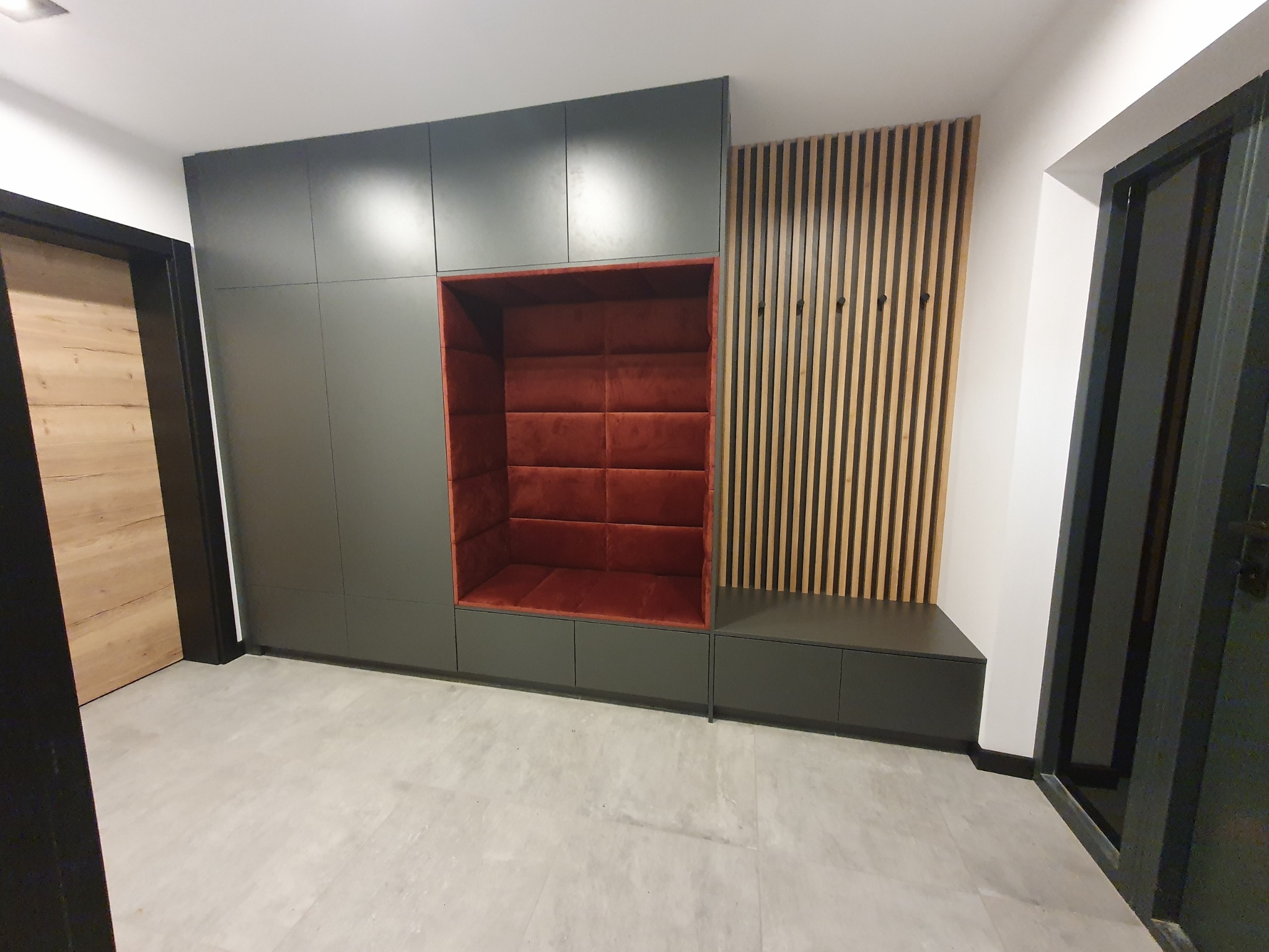 Wnęka tapicerowana w kolorze ceglastej czerwieni, wkomponowana w szarą zabudowę meblową w przedpokoju, obok ściana z drewnianych listew z haczykami, widok z dołu.