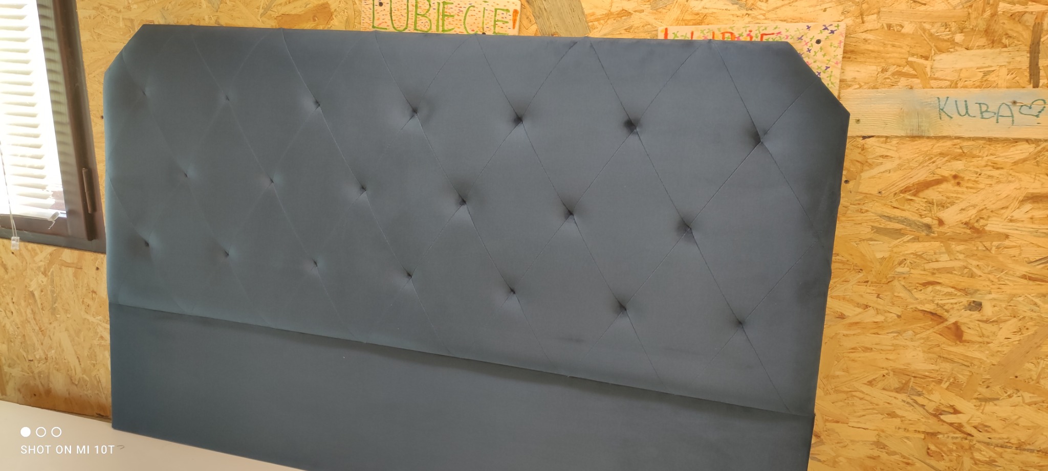 Szary, tapicerowany zagłówek łóżka z pikowaniem w romby, ustawiony na tle płyty OSB z napisem 'KUBA❤'.