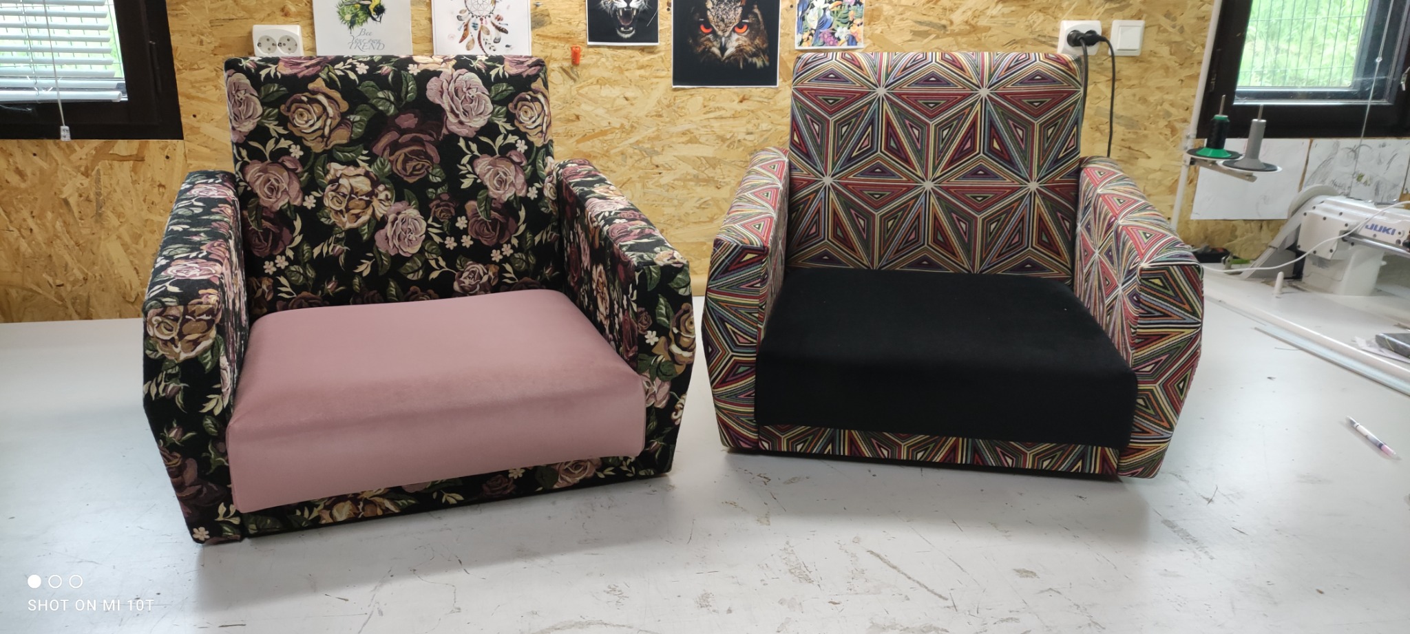 Dwa fotele po renowacji: jeden z obiciem w ciemne róże na czarnym tle i różowym siedziskiem, drugi w geometryczne wzory z czarnym siedziskiem, na tle warsztatu z maszynami do szycia.