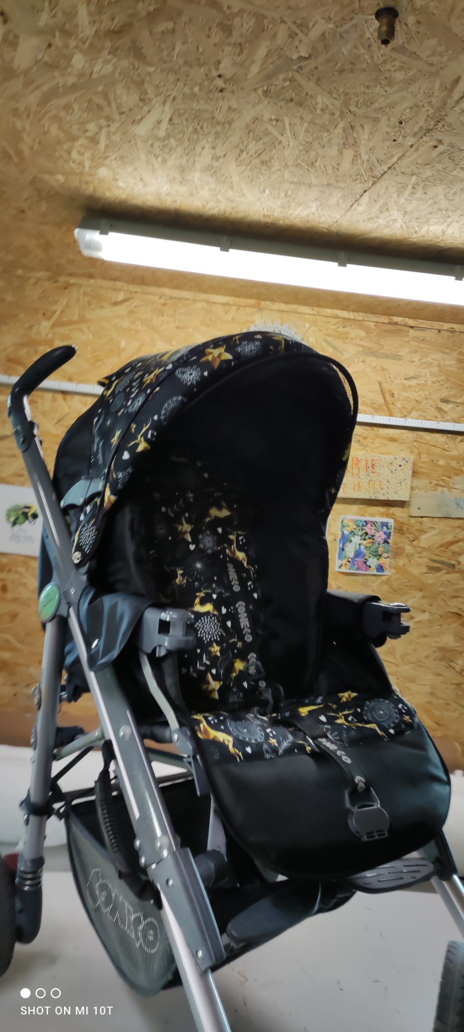 Wózek dziecięcy z czarną tapicerką w złote gwiazdy i białe wzory, sfotografowany w warsztacie z płyt OSB w tle, oświetlony lampą jarzeniową.