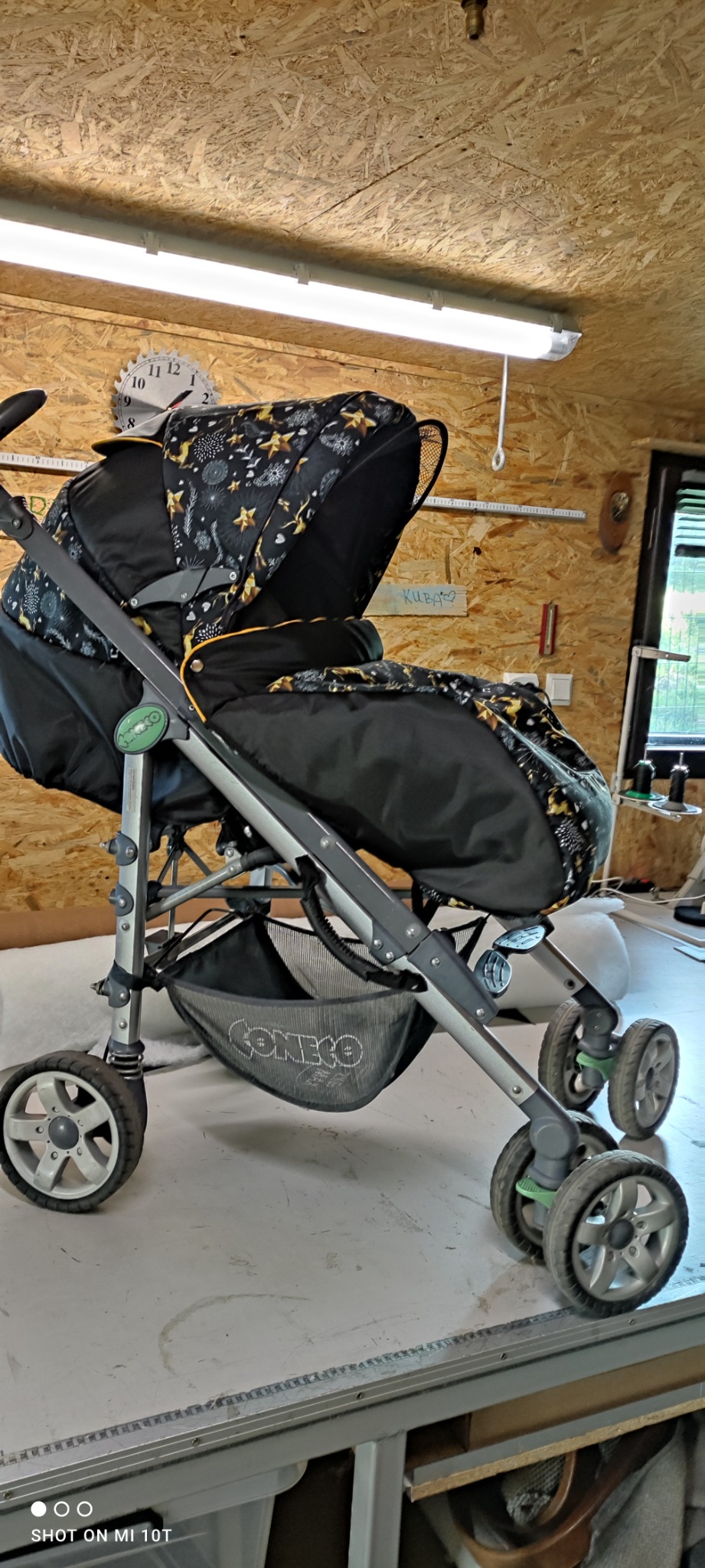 Wózek dziecięcy z nową, czarno-złotą tapicerką w warsztacie, widoczne ślady użytkowania na kołach i rama wózka.