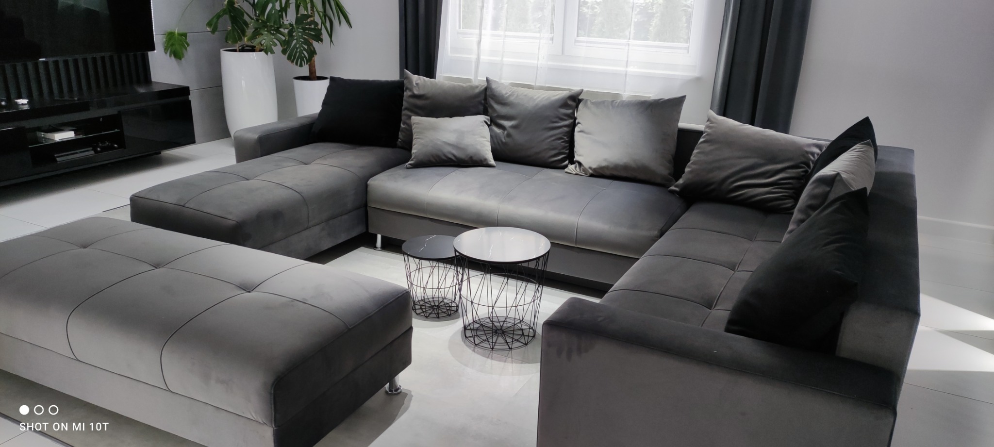 Szary, modułowy narożnik z pufą i dekoracyjnymi poduszkami w salonie, z dwoma okrągłymi stolikami kawowymi z metalową podstawą i marmurowym blatem.