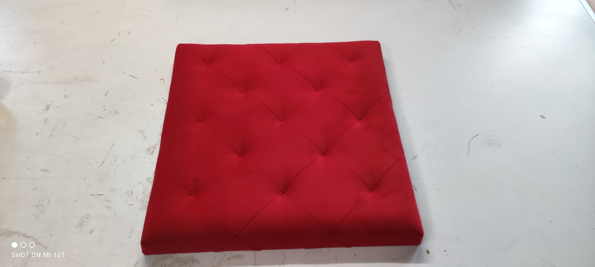 Czerwony, kwadratowy panel tapicerowany z regularnym wzorem pikowania na białym, lekko zabrudzonym tle.