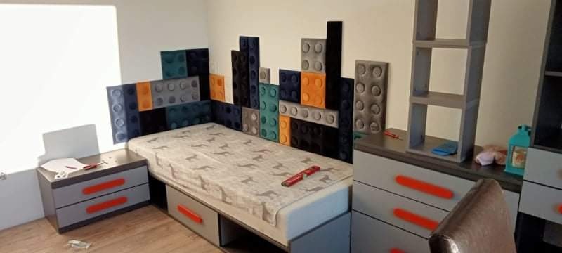 Pokój dziecięcy z łóżkiem i tapicerowanym zagłówkiem w kształcie klocków Lego, szary stolik nocny z pomarańczowymi uchwytami i regał na ścianie.