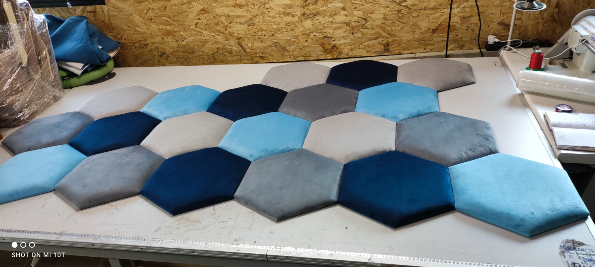 Układanka z sześciokątnych paneli tapicerowanych w odcieniach błękitu, szarości i granatu, ułożonych na stole w warsztacie, widoczna maszyna do szycia i akcesoria.