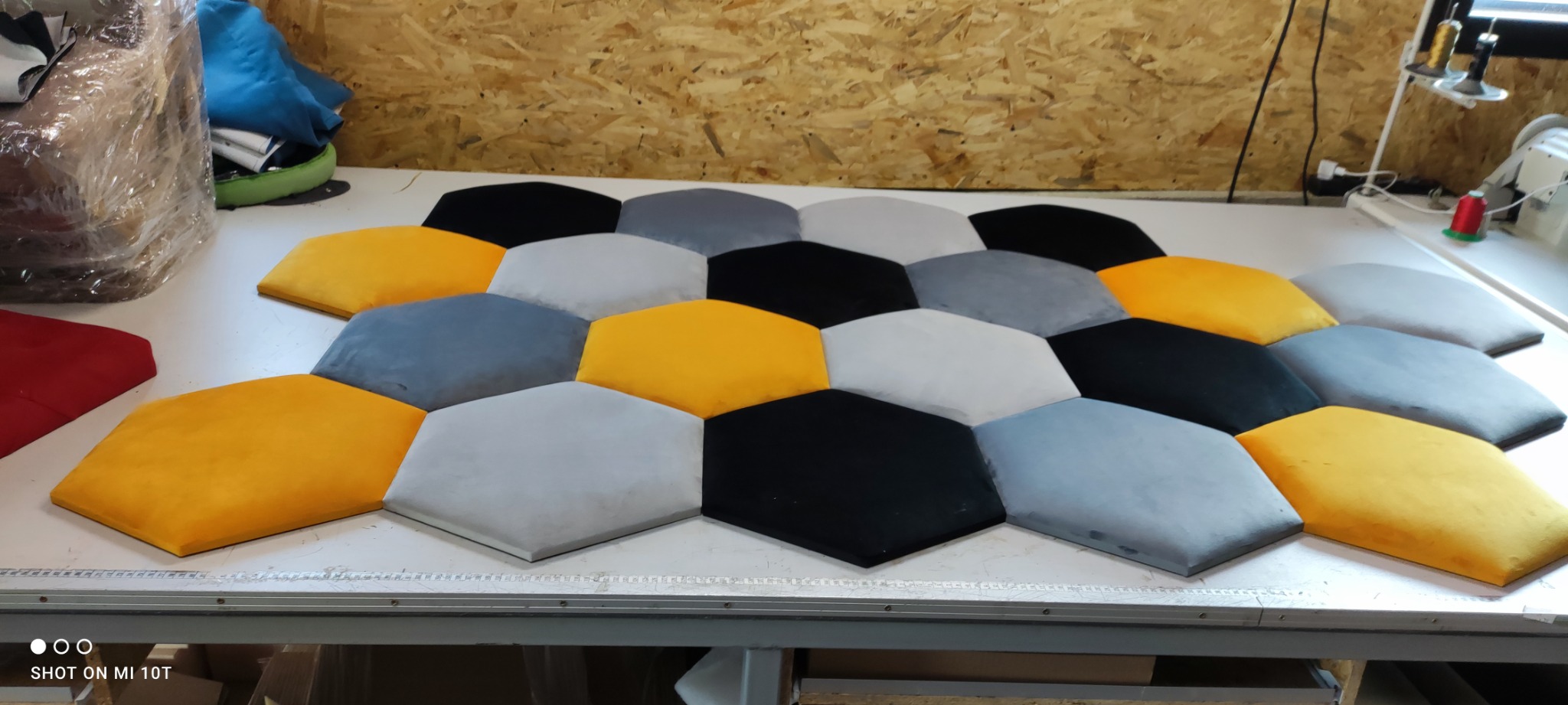 Wielobarwne, sześciokątne panele tapicerowane (żółte, szare, czarne) ułożone na stole w warsztacie, widoczne narzędzia krawieckie w tle.