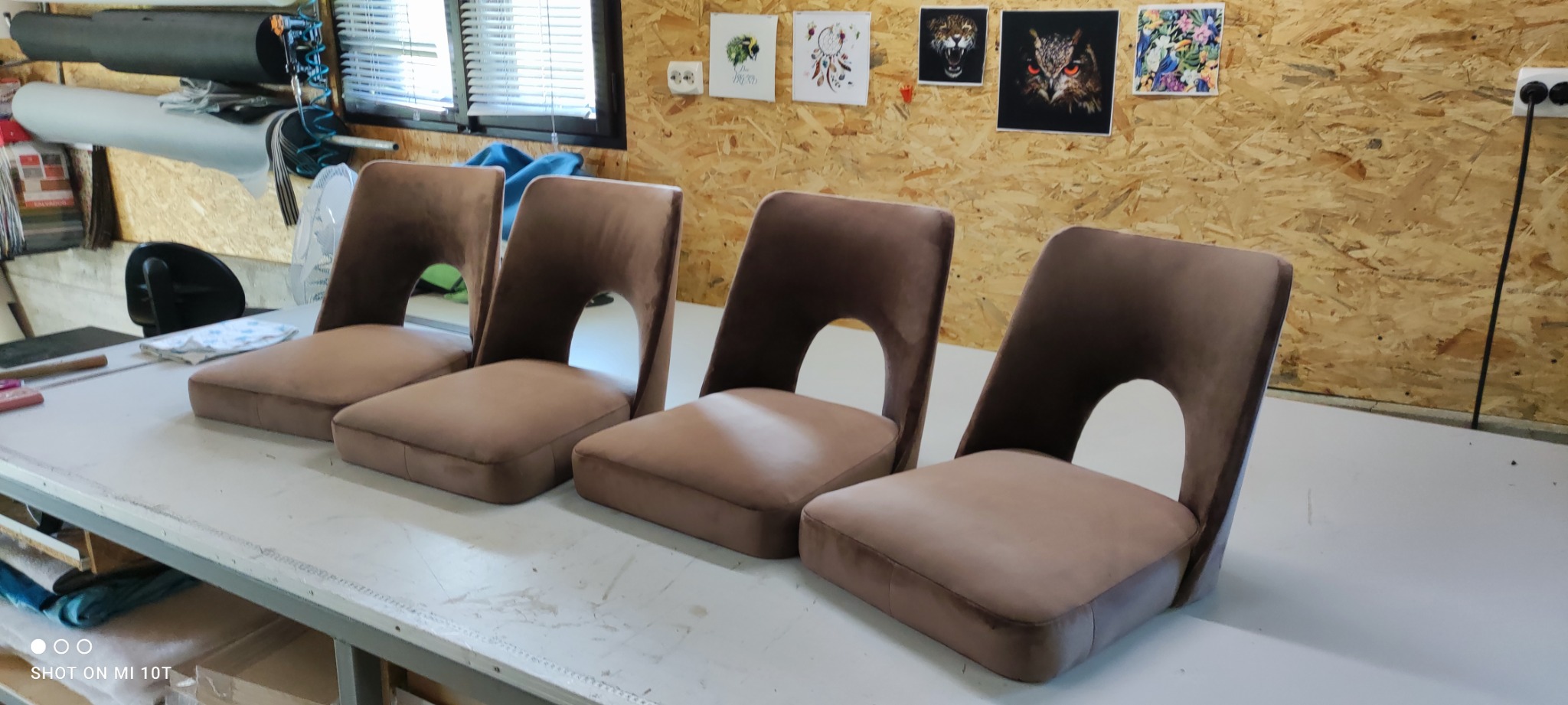 Cztery krzesła z brązową, welurową tapicerką, ustawione w rzędzie na stole w warsztacie tapicerskim. W tle widoczna ściana z płyt OSB i grafiki na ścianie.