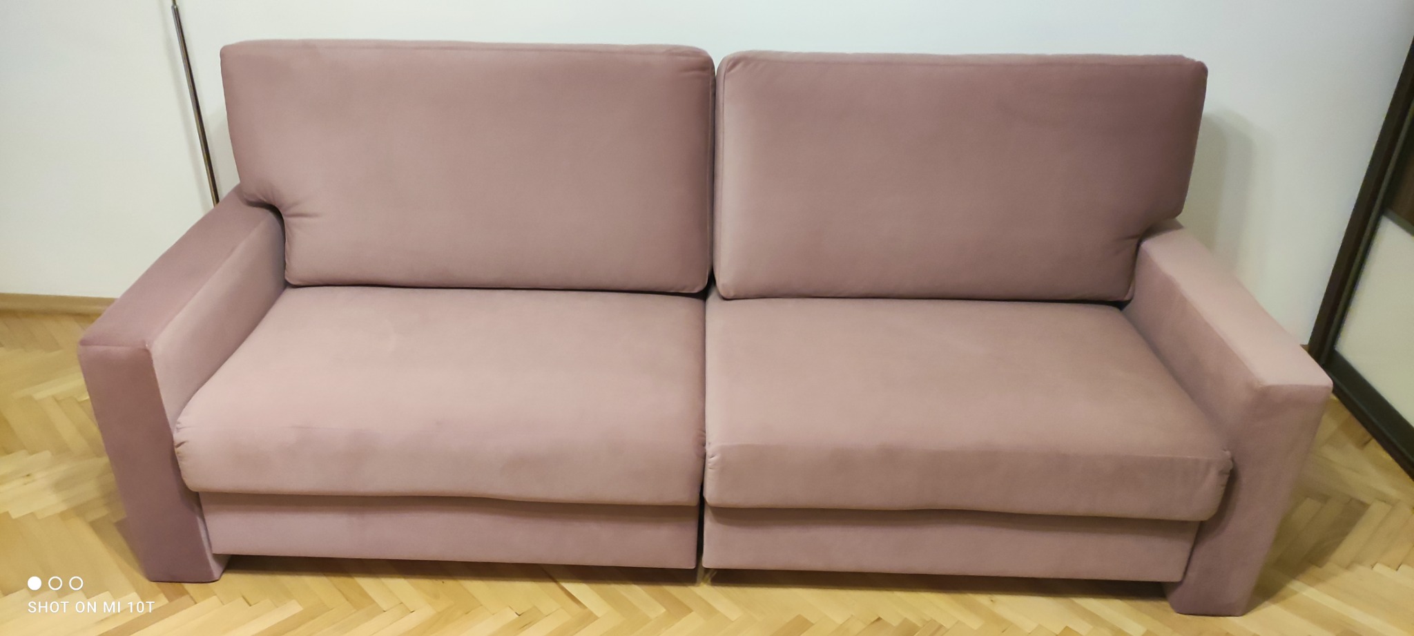 Dwuosobowa sofa w odcieniu pudrowego różu z welurową tapicerką, widoczna na tle parkietu i białej ściany, z delikatnym odblaskiem światła na siedzisku.