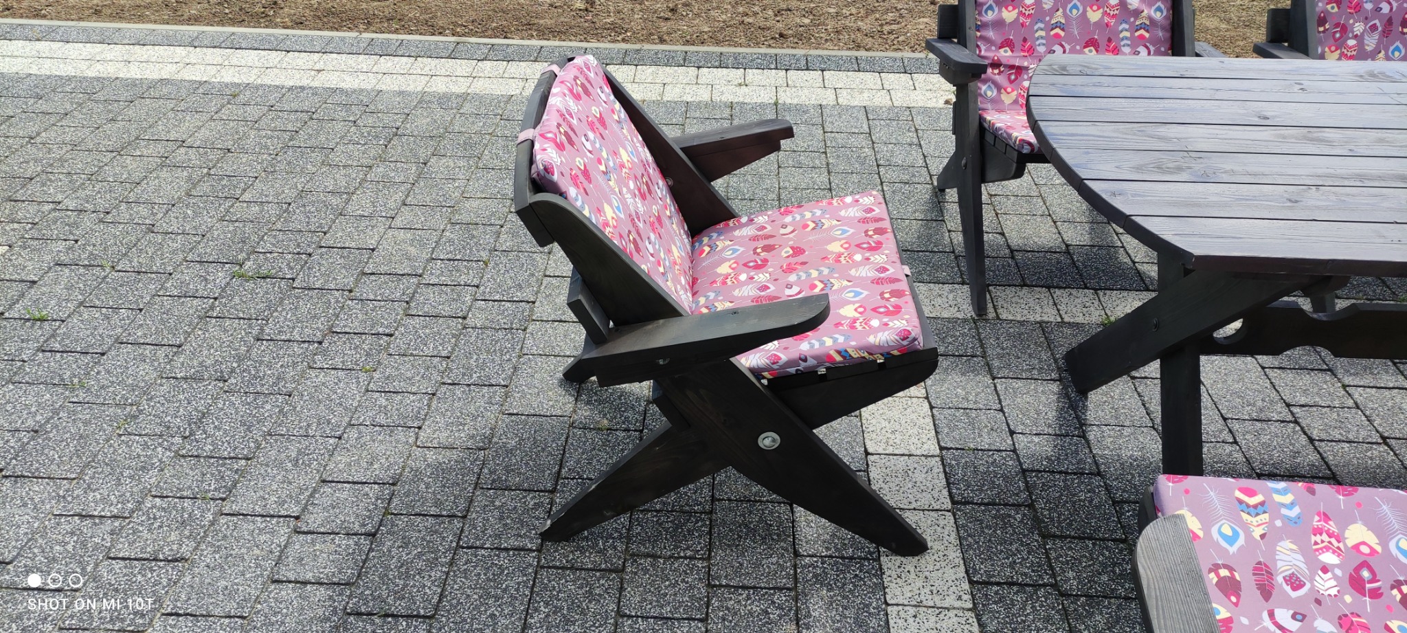 Ciemne drewniane meble ogrodowe z poduszkami w motywie piór na szarym brukowanym patio.