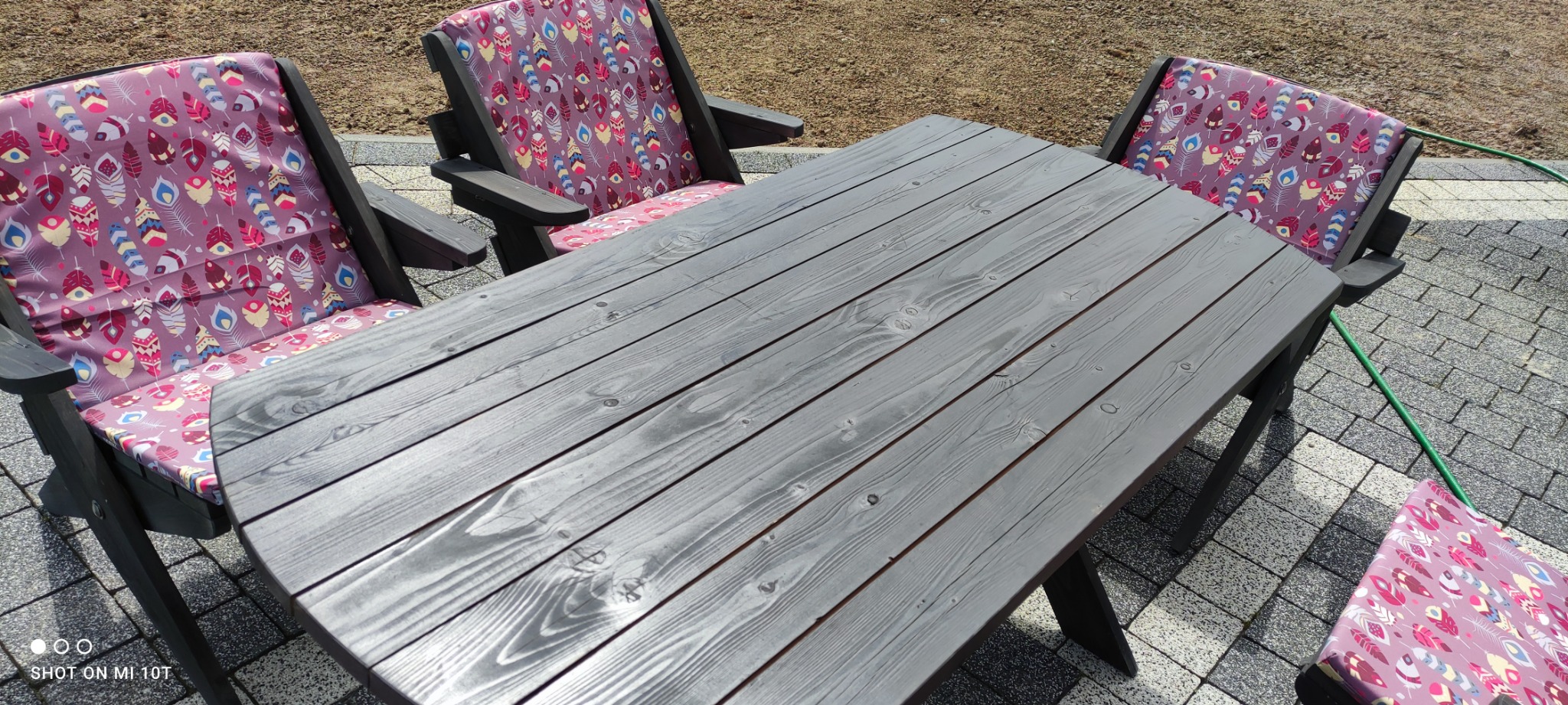 Drewniany stół i krzesła ogrodowe z poduszkami w piórka, ustawione na kostce brukowej, widoczne ślady malowania szarą farbą.