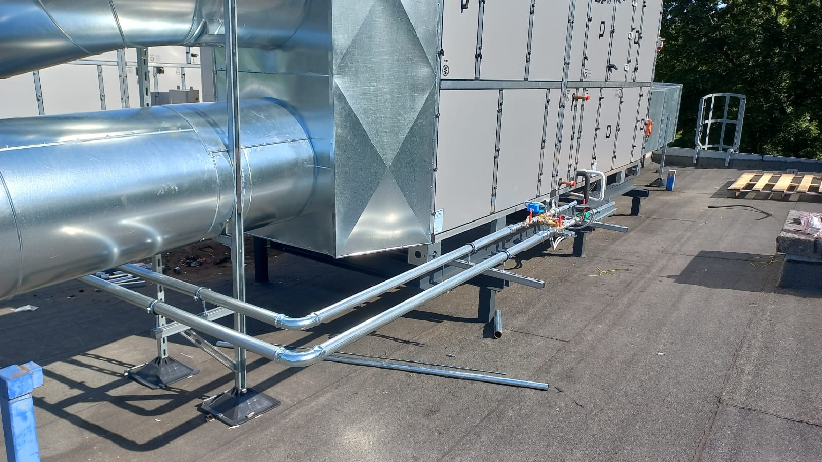 Instalacja przemysłowej klimatyzacji na dachu budynku, widoczne duże kanały wentylacyjne, rury chłodnicze z zaworami, elementy konstrukcyjne wsporcze i fragment zieleni w tle.