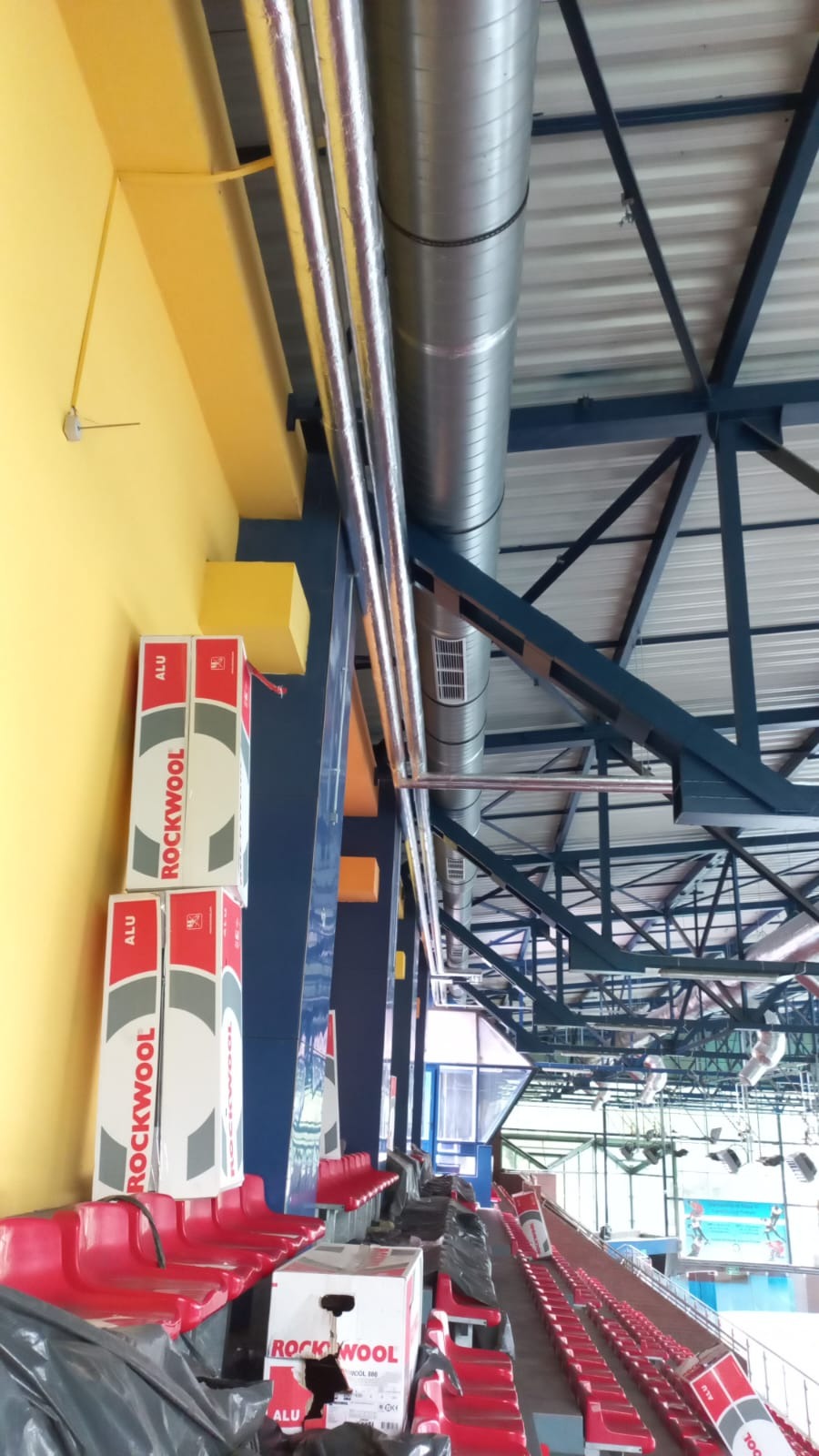 Wysoko zamontowane, ocieplone rury wentylacyjne w hali sportowej, widoczne pakiety wełny mineralnej Rockwool ALU, żółto-niebieska konstrukcja nośna i rząd czerwonych siedzeń.