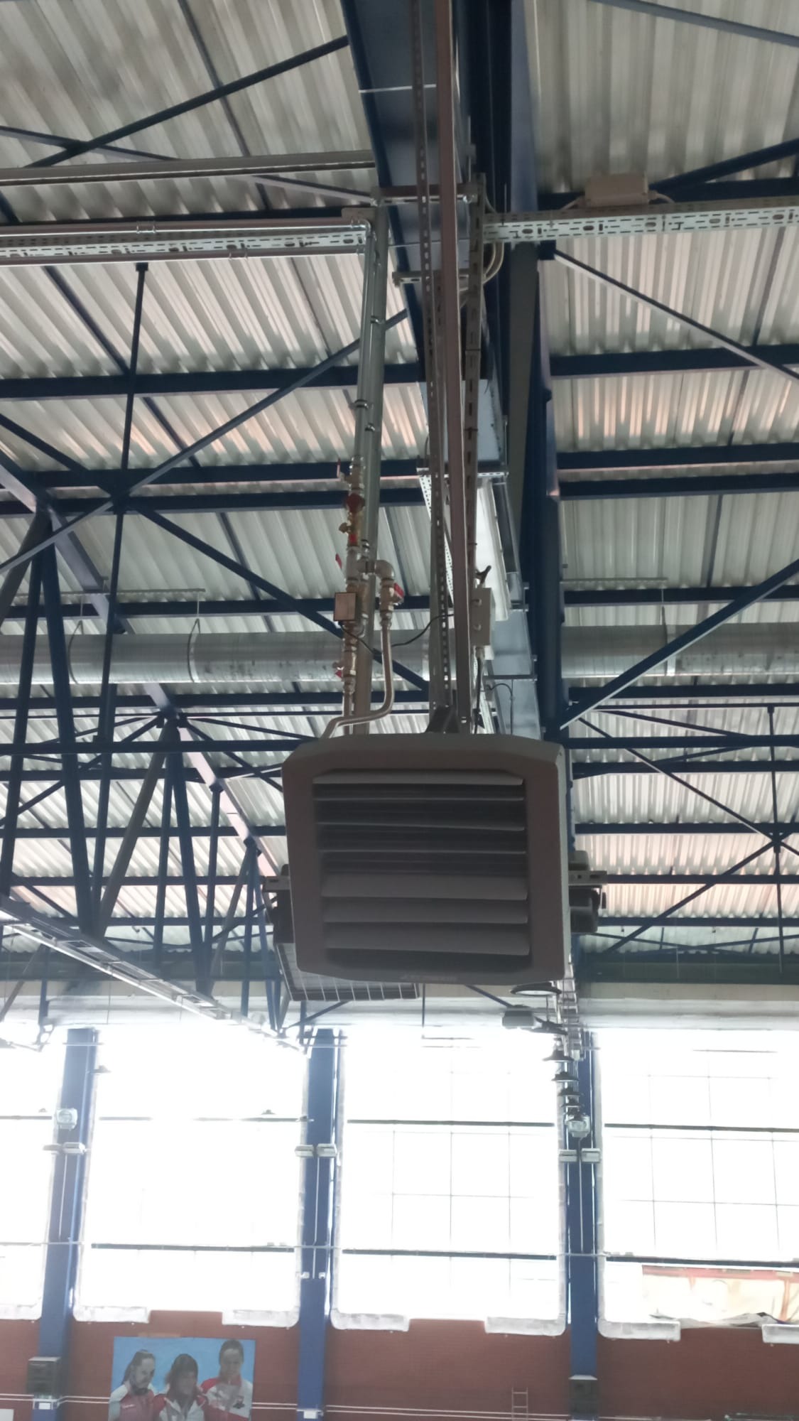 Sufitowa nagrzewnica powietrza z podłączonymi rurami w hali sportowej, widok z dołu na tle metalowej konstrukcji dachu i okien.