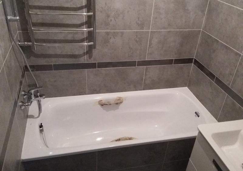 Biała wanna obudowana szarymi płytkami z ciemniejszym pasem, bateria wannowa z prysznicem, grzejnik drabinkowy na ścianie, widoczne ślady kleju po uszczelnieniu wanny.