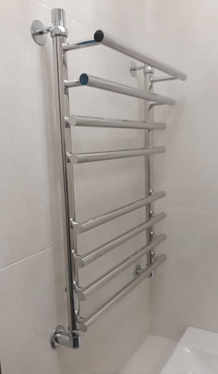 Chromowany, drabinkowy grzejnik łazienkowy zamontowany na białej, kafelkowanej ścianie, widoczne elementy mocujące.