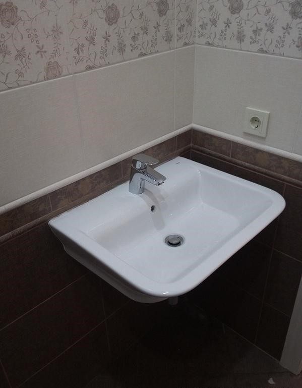 Biała umywalka z chromowaną baterią zamontowana w narożniku łazienki, z brązowymi i beżowymi płytkami, na ścianie widoczne gniazdko elektryczne.