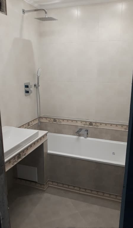 Wykończona łazienka z białą wanną, umywalką i deszczownicą. Beżowe płytki na ścianach i podłodze, dekoracyjne listwy przy wannie i umywalce.