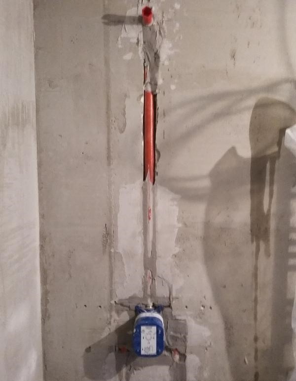 Szara ściana w trakcie prac hydraulicznych z widoczną czerwoną rurą w bruździe i niebieskim zaworem podtynkowym.