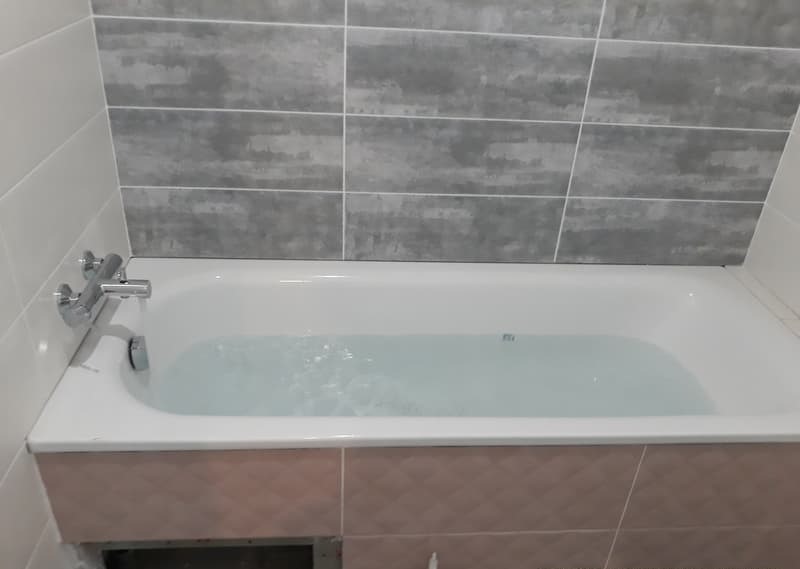 Napełniona wodą biała wanna z chromowaną baterią na tle szaro-białych płytek w łazience.