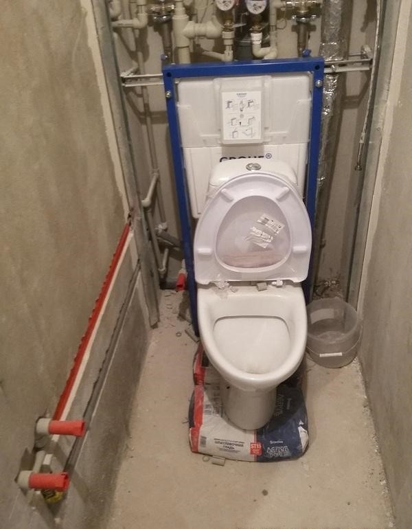 Biała toaleta wisząca Grohe w trakcie montażu, ustawiona na worku zaprawy, widoczne rury instalacyjne w surowym wnętrzu łazienki.