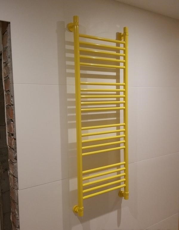 Żółty grzejnik łazienkowy drabinkowy zamontowany na białej, kafelkowanej ścianie. Widoczny fragment ściany z surową cegłą.