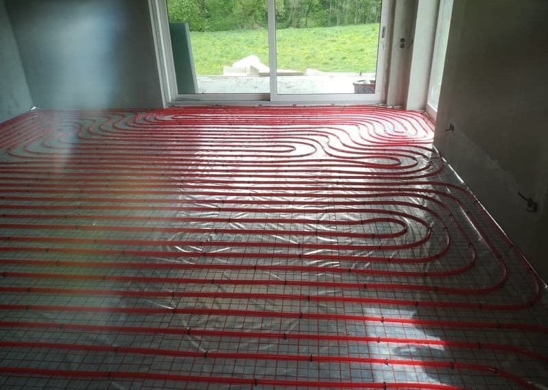 Czerwone rury ogrzewania podłogowego ułożone na siatce, pokryte folią, w surowym wnętrzu z oknem na zieleń.
