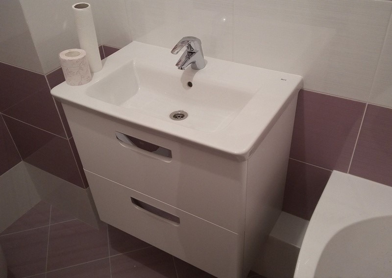 Biała szafka pod umywalkę z dwoma szufladami, chromowana bateria, rolka papieru toaletowego i zapasowa rolka na blacie, w tle biało-fioletowe płytki.